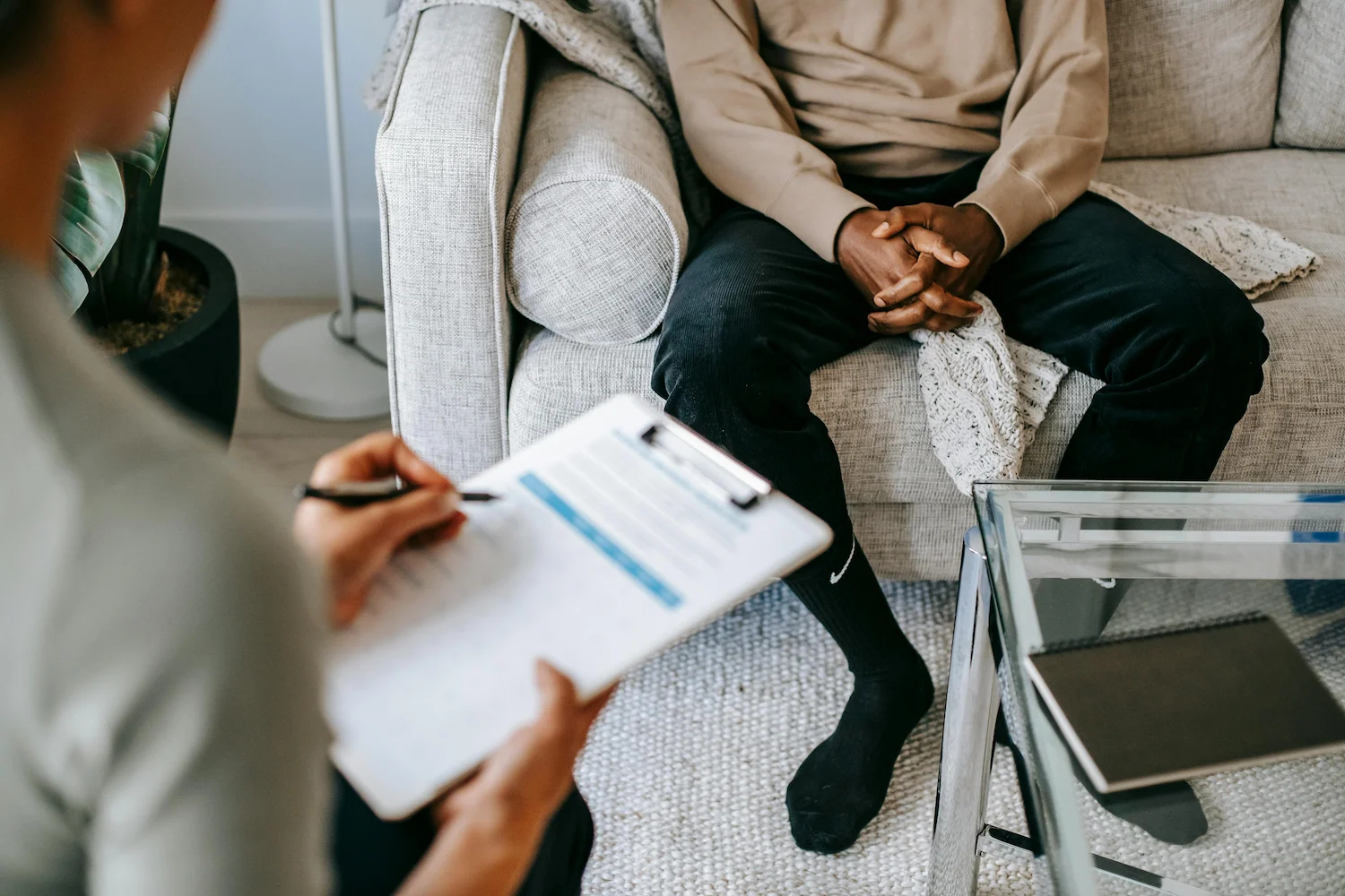 Black male sitting on a couch talking with a therapist holding a clipboard.
