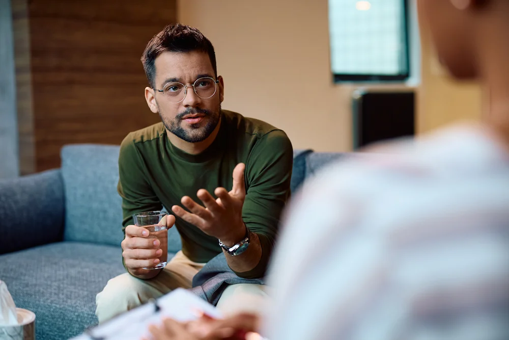 Male with glasses sitting on a couch talking with a therapist.